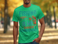 A fishing enthusiast sports a Garment Graphics green t-shirt emblazoned with Fishing Is My Anger Management and decorated with fish hooks and a fish, set against blurry trees in the background.