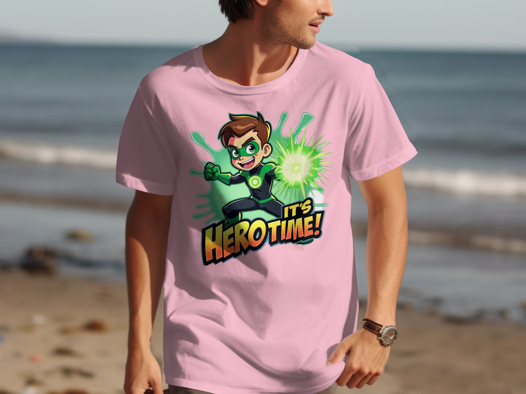 Someone wearing a vibrant pink Garment Graphics T-shirt with a superhero cartoon holding a glowing green orb and the text Its Hero Time! stands on the beach, the ocean providing the perfect backdrop for this fun family adventure.
