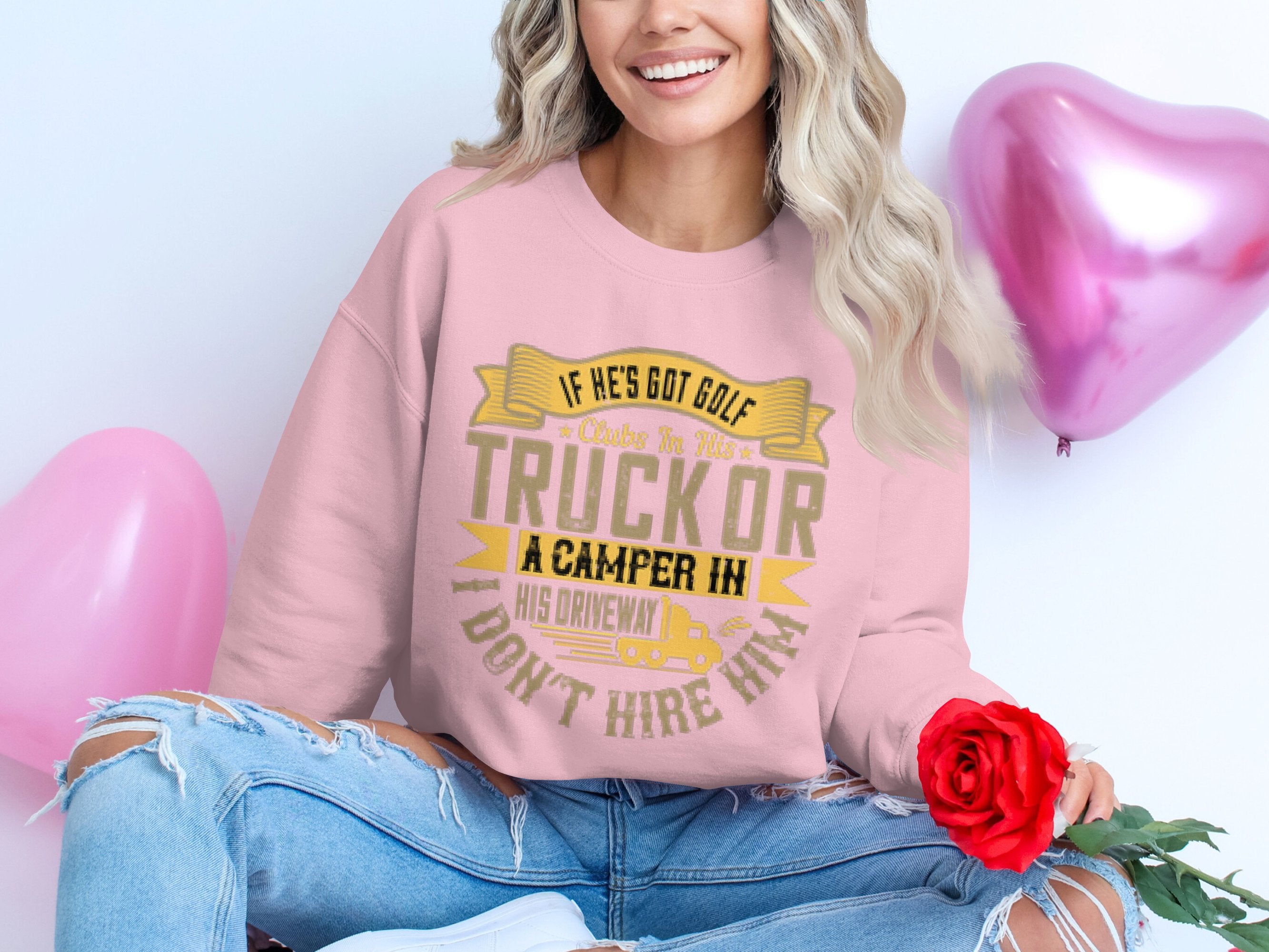A woman sits cross-legged on the floor, holding a red rose, wearing Garment Graphics pink sweatshirt with amusing golf trucks and campers text. Her ripped jeans match her cheerful smile, while two heart-shaped balloons enhance the playful ambiance.