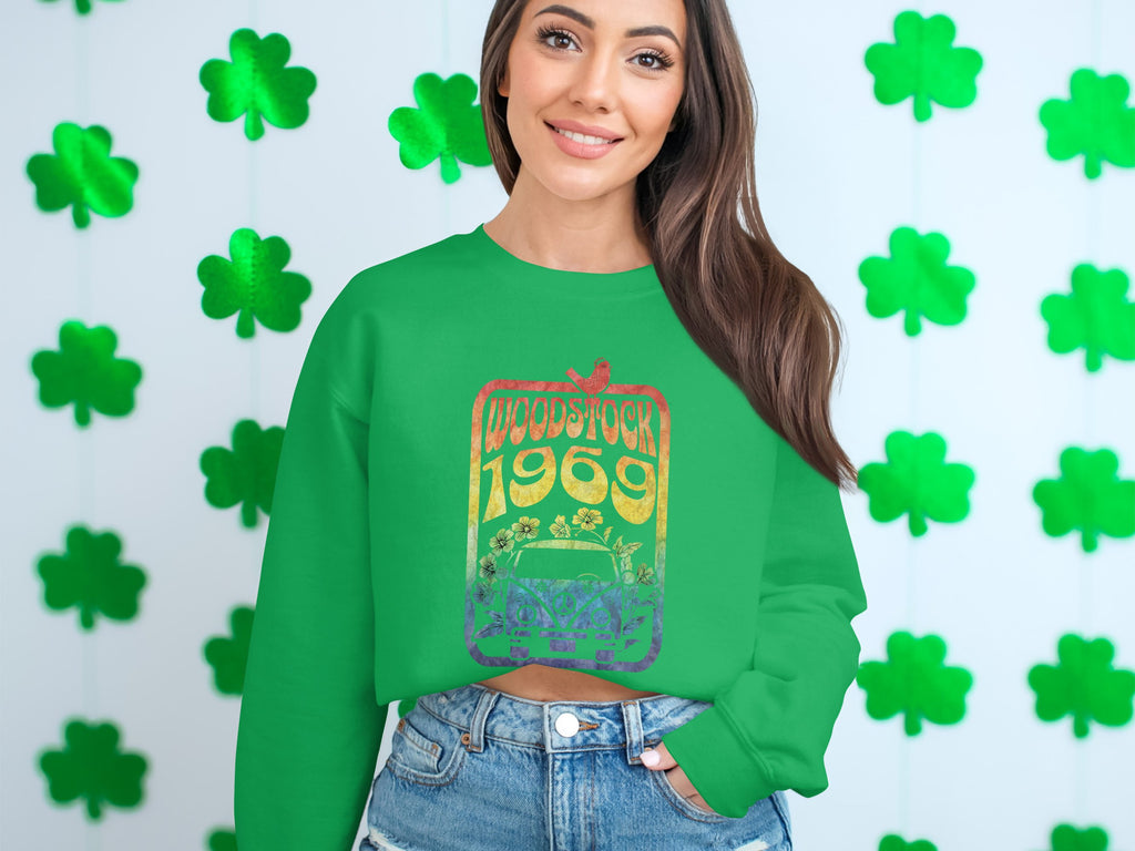 A person sports a green Garment Graphics sweatshirt featuring a Woodstock 1969 design, standing before green shamrock decor for a retro look. They have long dark hair and wear denim shorts.