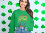 A person sports a green Garment Graphics sweatshirt featuring a Woodstock 1969 design, standing before green shamrock decor for a retro look. They have long dark hair and wear denim shorts.