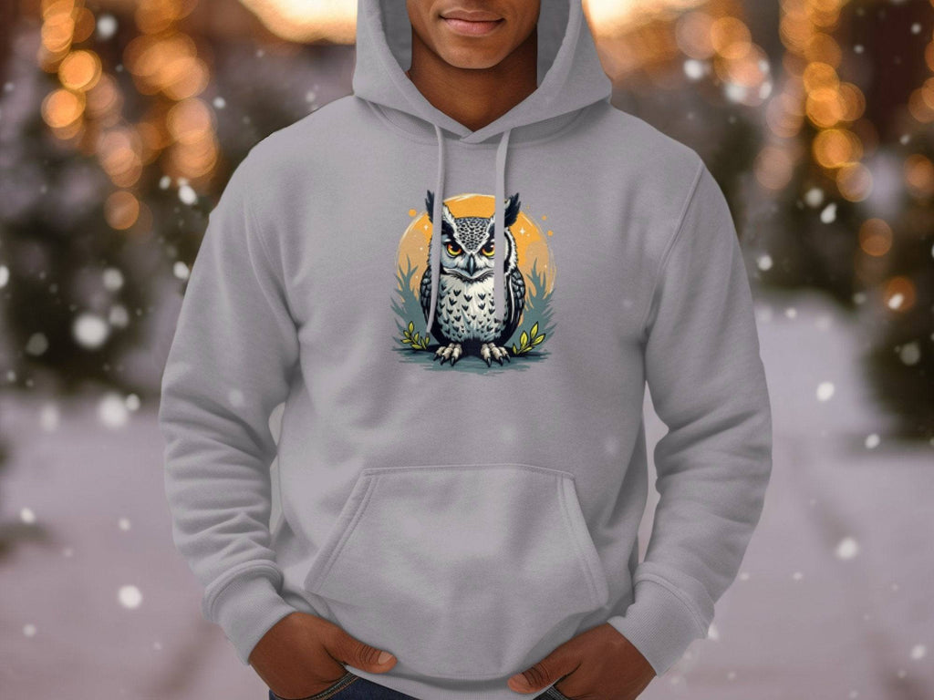 A person wears a classic fit hoodie from Garment Graphics, featuring a detailed owl graphic on a branch. The medium-heavy fabric complements the blurred backdrop with holiday lights and falling snow, creating a cozy atmosphere.