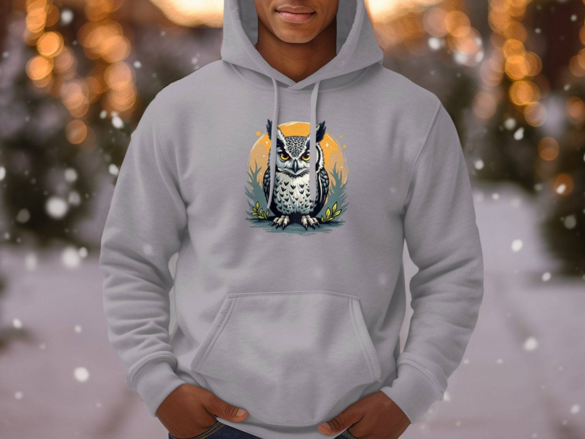 A person wears a classic fit hoodie from Garment Graphics, featuring a detailed owl graphic on a branch. The medium-heavy fabric complements the blurred backdrop with holiday lights and falling snow, creating a cozy atmosphere.