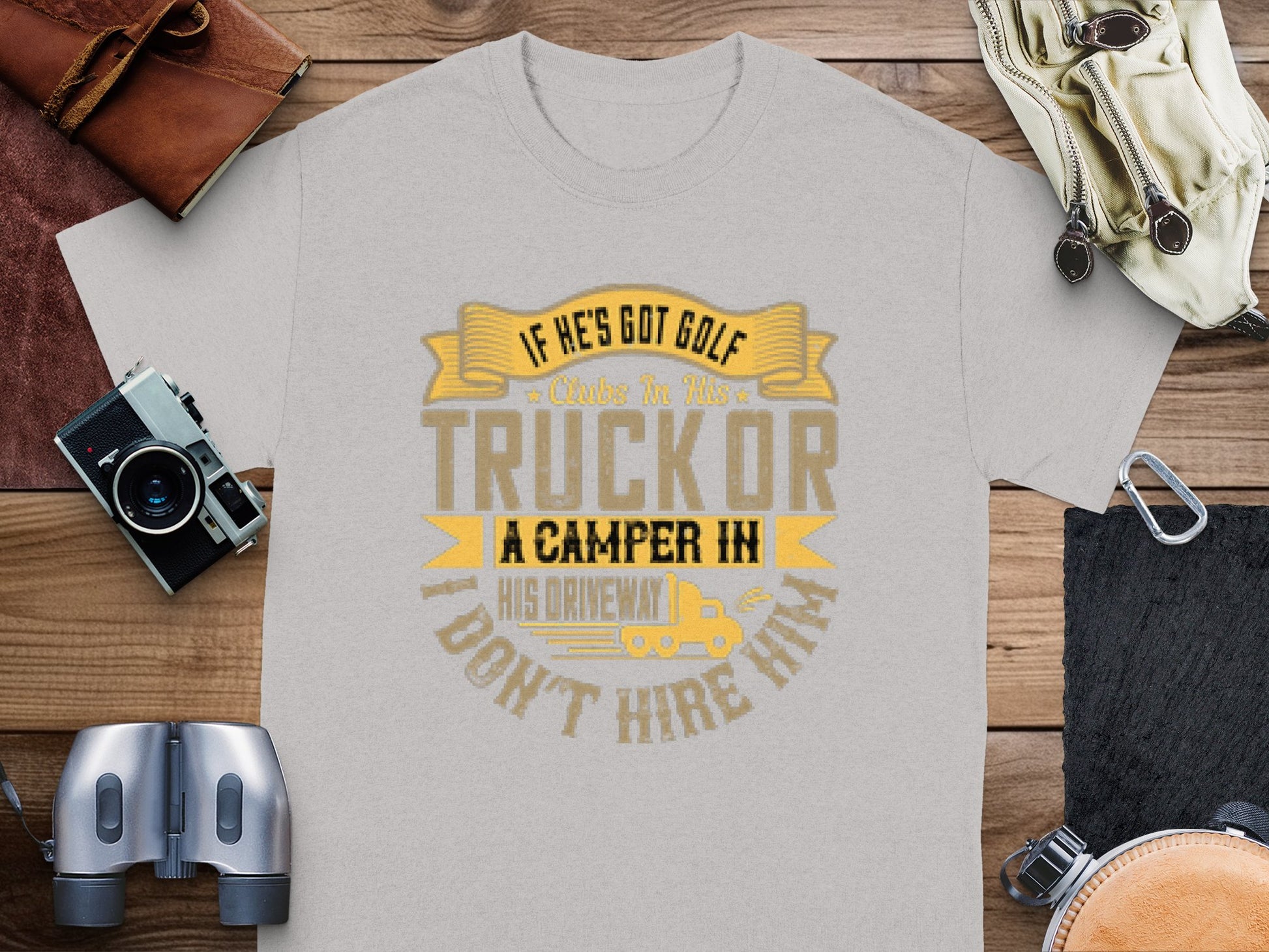 A Garment Graphics gray t-shirt with the text If hes got golf clubs in his truck or a camper in his driveway, dont hire him sits on a wooden surface, surrounded by camping essentials: a camera, wallet, binoculars, and papers.