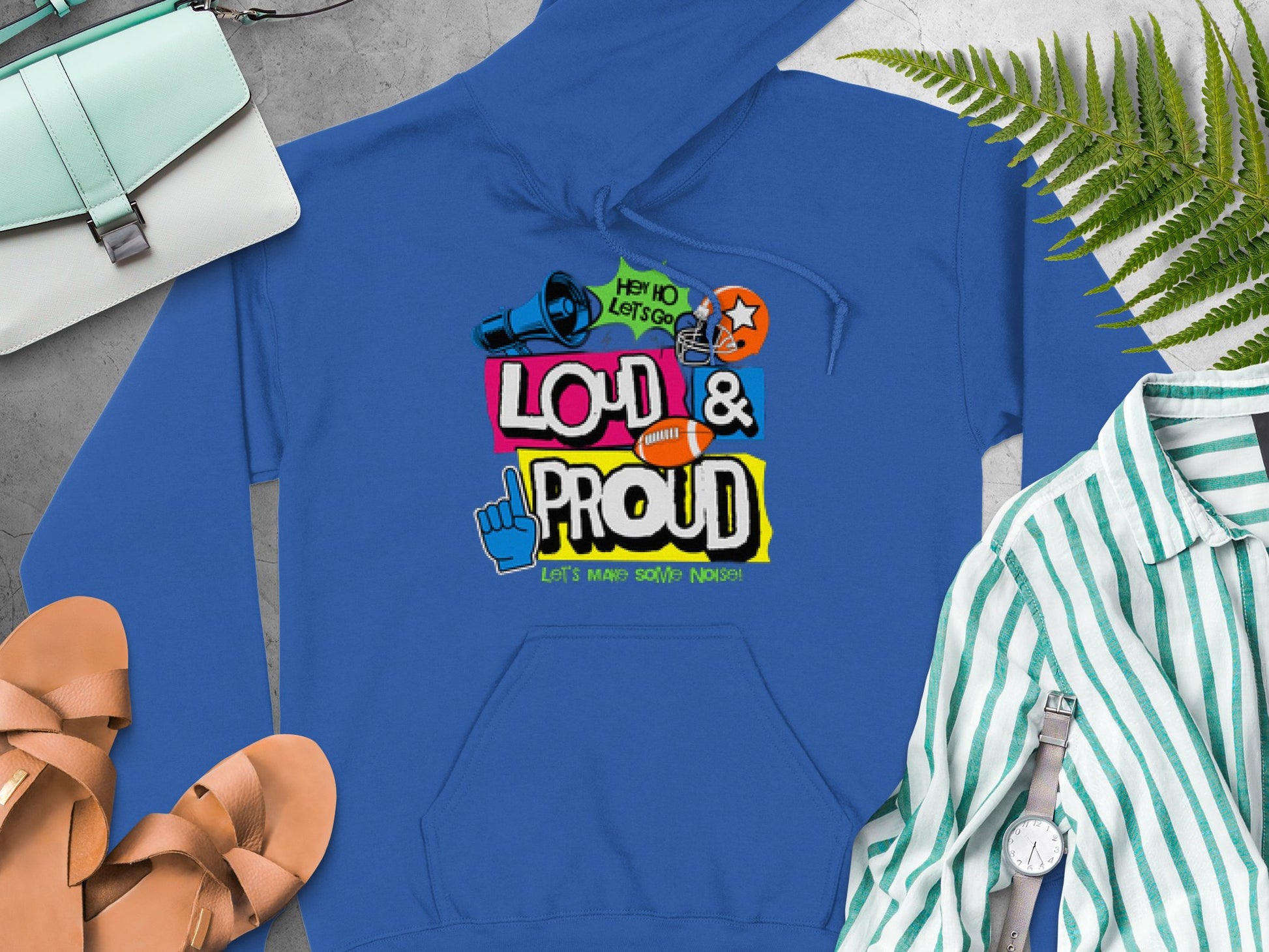 A blue hoodie from Garment Graphics features vibrant Loud & Proud text, a megaphone, basketball, and peace sign graphics. Its styled with a purse, sandals, and a striped shirt on a gray surface accented by green fern leaves.