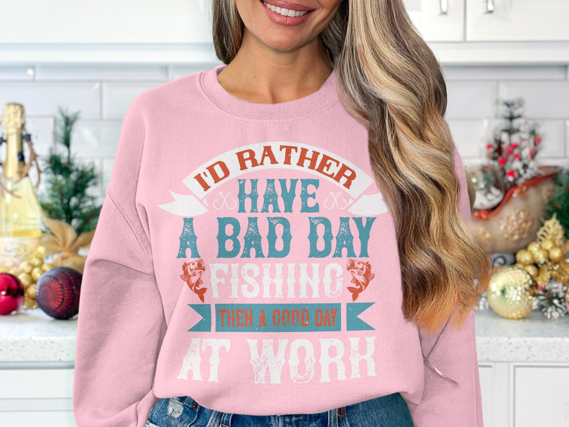 In her kitchen filled with holiday decorations, a woman wears a pink sweatshirt from Garment Graphics Bad Day Fishing Sweatshirts collection that reads, Id rather have a bad day fishing than a good day at work.