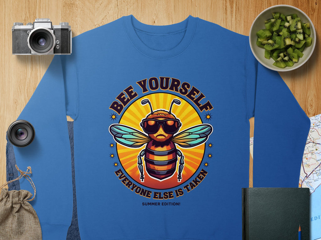 Garment Graphics offers a blue unisex sweatshirt featuring a bee with sunglasses and Bee Yourself, Everyone Else Is Taken, Summer Edition! text. The colorful design appears alongside a camera, lens, chopped cucumbers in a bowl, and notebook on textured fabric.