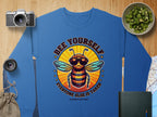 Garment Graphics offers a blue unisex sweatshirt featuring a bee with sunglasses and Bee Yourself, Everyone Else Is Taken, Summer Edition! text. The colorful design appears alongside a camera, lens, chopped cucumbers in a bowl, and notebook on textured fabric.