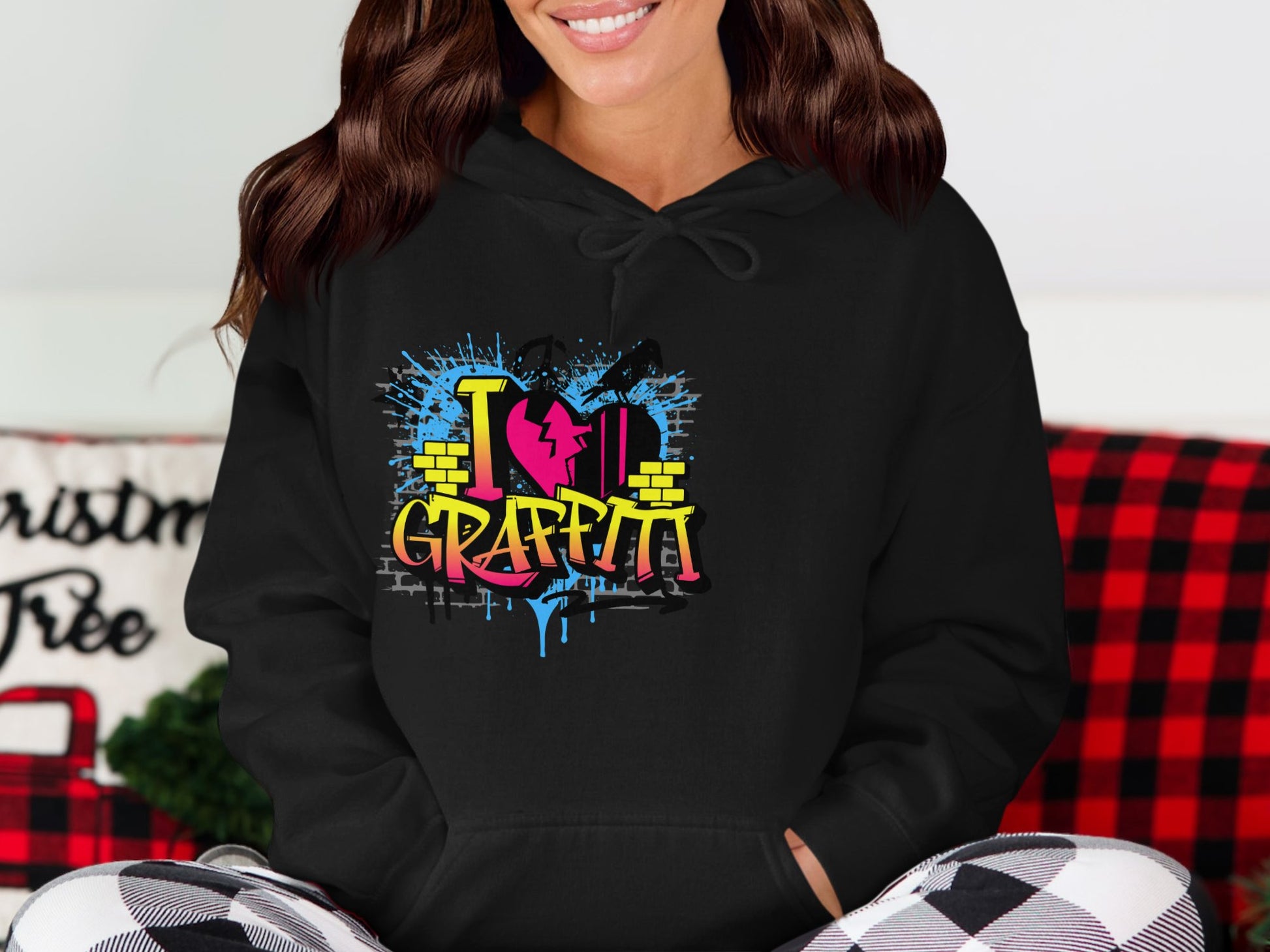 A person sports a Garment Graphics black hoodie with I Love Graffiti in colorful graffiti text, featuring a brick wall and heart motif. They sit before a festive backdrop on a red and black checkered blanket, ideal for any casual urban street art-inspired wardrobe.