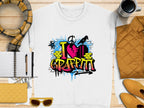 A Garment Graphics urban style white T-shirt displays a vibrant graffiti design with I Love Graffiti, brick patterns, and paint splatters. Its arranged alongside a scarf, notebook, pen, coffee cup, quilted items, and sunglasses.