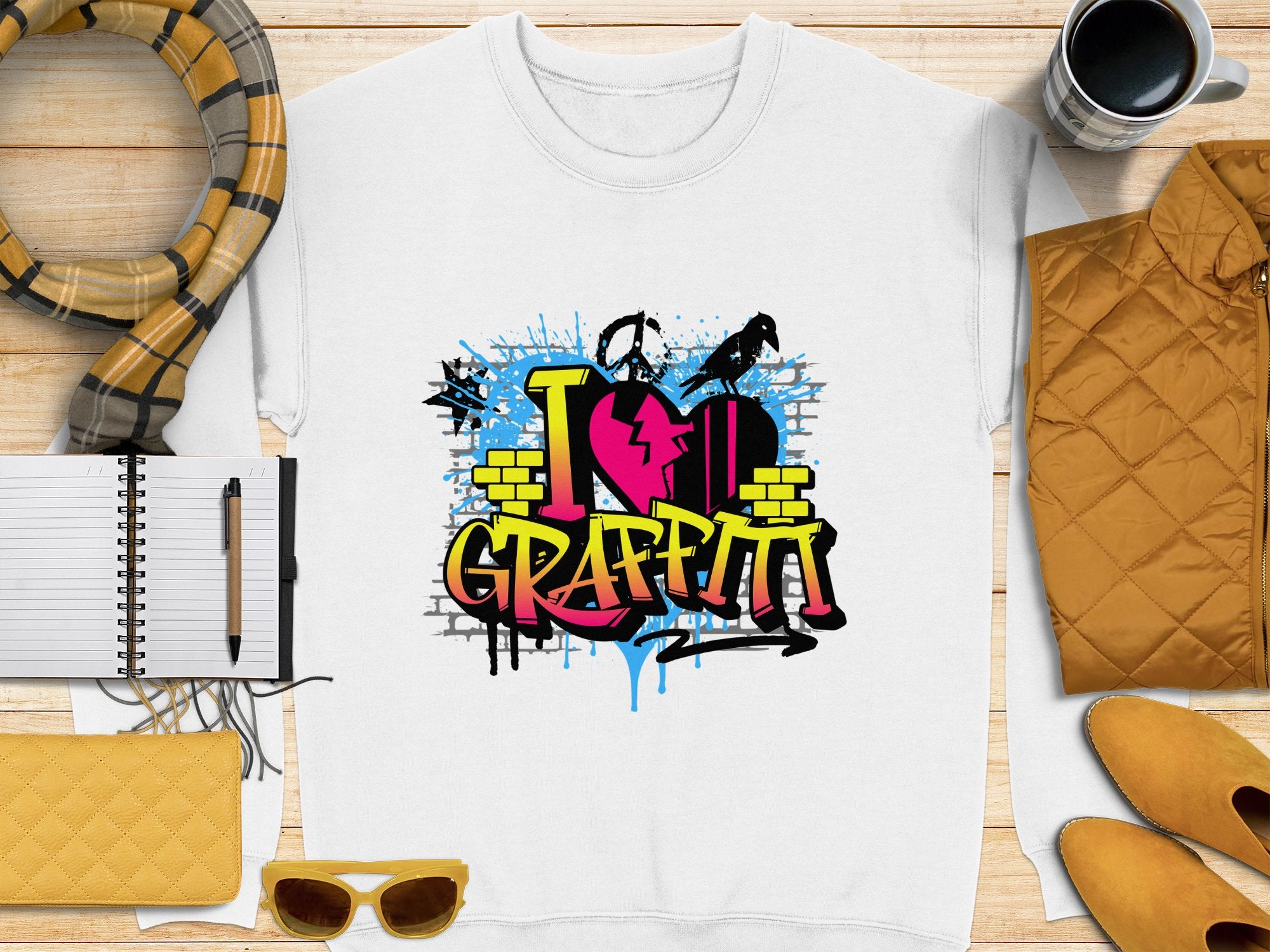 A Garment Graphics urban style white T-shirt displays a vibrant graffiti design with I Love Graffiti, brick patterns, and paint splatters. Its arranged alongside a scarf, notebook, pen, coffee cup, quilted items, and sunglasses.