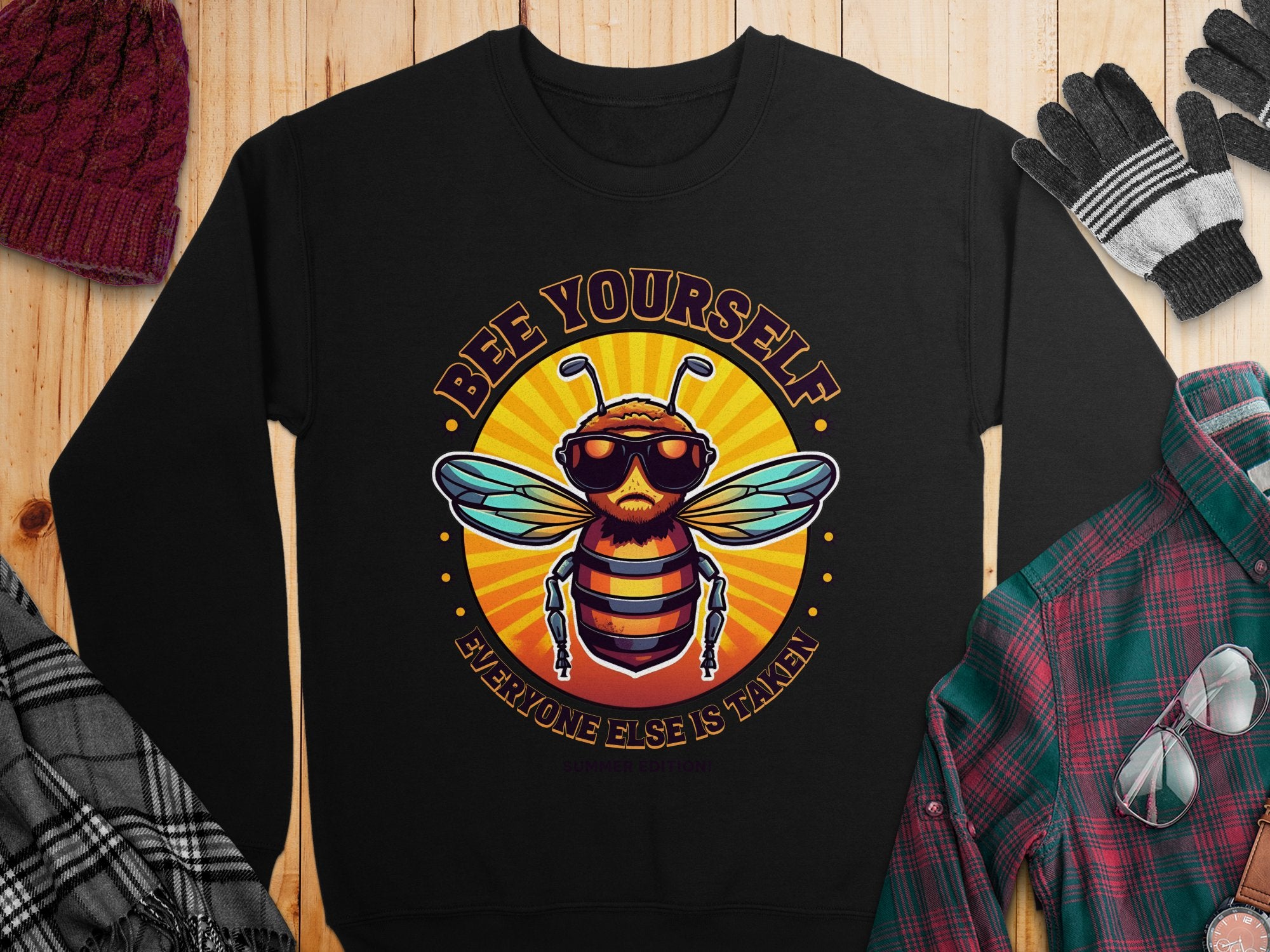 A black, unisex sweatshirt from Garment Graphics lies on wood, featuring a cartoon bee with sunglasses and the phrase Bee Yourself, Everyone Else is Taken. Nearby are a plaid shirt, scarf, gloves, and hat. Ideal for breezy summer nights.