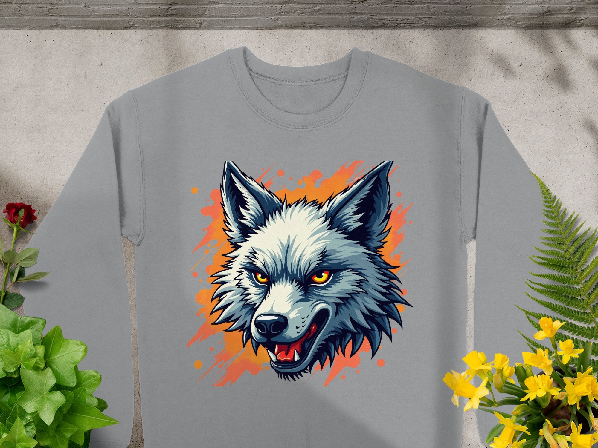 Garment Graphics presents a gray sweatshirt adorned with a fierce wolf graphic, showcasing sharp teeth and striking yellow eyes against an orange splash. Surrounded by green plants and yellow flowers on concrete, its a bold statement piece capturing the wild side.