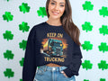 A woman with long hair smiles at the camera, showcasing a Garment Graphics sweatshirt perfect for truck lovers. Her navy top sports a truck and Keep on Trucking. She stands against a white wall decorated with green shamrock cutouts, paired with light blue jeans.