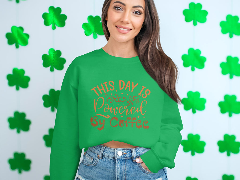A woman with long brown hair smiles, wearing a Garment Graphics sweatshirt that displays This day is powered by coffee in orange. She stands against a white wall decorated with green four-leaf clovers.