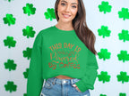 A woman with long brown hair smiles, wearing a Garment Graphics sweatshirt that displays This day is powered by coffee in orange. She stands against a white wall decorated with green four-leaf clovers.