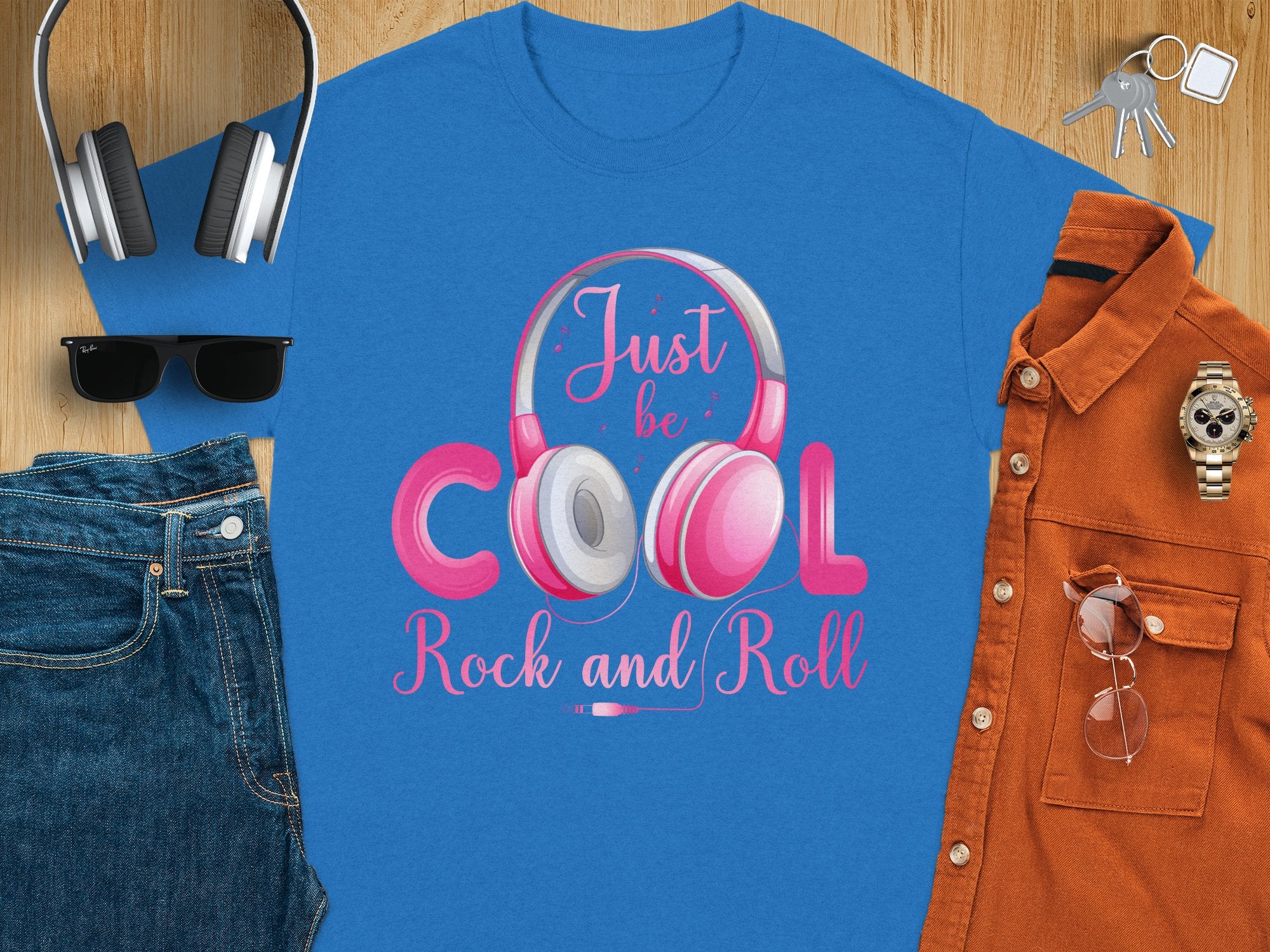 A flat lay for music lovers includes a blue Just be cool, rock and roll t-shirt by Garment Graphics with pink and white headphones. Also featured are sunglasses, keys, blue jeans, an orange shirt, a wristwatch, and glasses—ideal for rock and roll t-shirt fans.