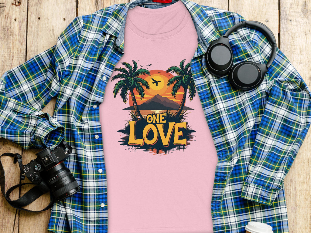 A pink Garment Graphics T-shirt featuring the One Love tropical sunset with palm trees and birds is displayed on a wooden surface. Ideal for the beach, its accompanied by a plaid shirt, camera, headphones, and a lidded cup.