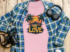 A pink Garment Graphics T-shirt featuring the One Love tropical sunset with palm trees and birds is displayed on a wooden surface. Ideal for the beach, its accompanied by a plaid shirt, camera, headphones, and a lidded cup.