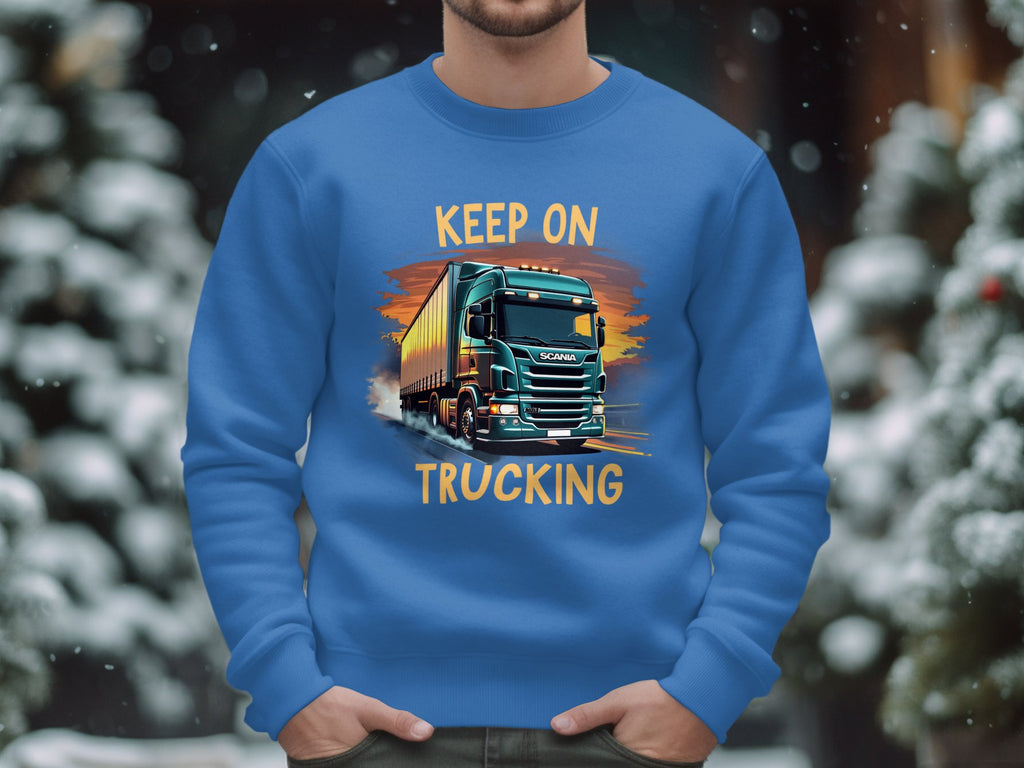 A person in a Garment Graphics sweatshirt with a bold truck design and Keep On Trucking stands against a snowy, out-of-focus tree backdrop, implying a winter scene.