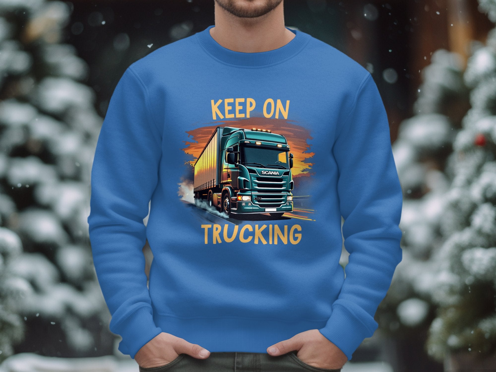 A person in a Garment Graphics sweatshirt with a bold truck design and Keep On Trucking stands against a snowy, out-of-focus tree backdrop, implying a winter scene.