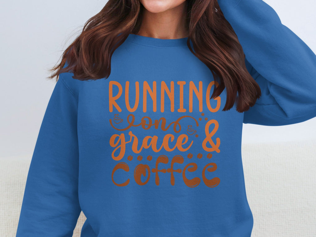 A person is wearing a Garment Graphics blue sweatshirt featuring the phrase Running on Grace and Coffee in orange. Known for its comfy unisex fit, it perfectly complements their long brown hair, though their face isnt visible in the frame.