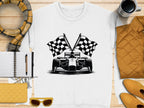 A white T-shirt by Garment Graphics, showcasing a checkered flag print and race car illustration, is displayed on a wooden surface. Accompanying items include a scarf, notebook, pen, yellow clutch, sunglasses, mustard jacket, and coffee cup.