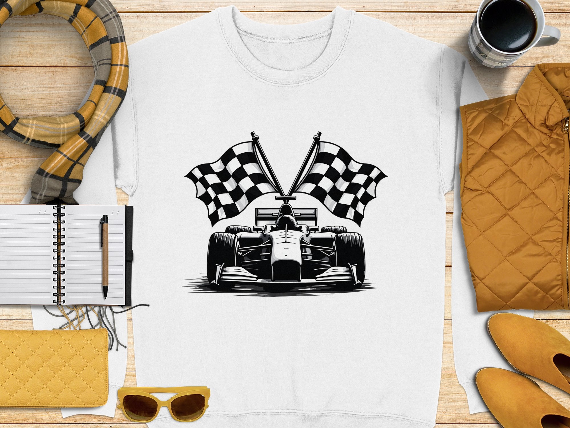 A white T-shirt by Garment Graphics, showcasing a checkered flag print and race car illustration, is displayed on a wooden surface. Accompanying items include a scarf, notebook, pen, yellow clutch, sunglasses, mustard jacket, and coffee cup.