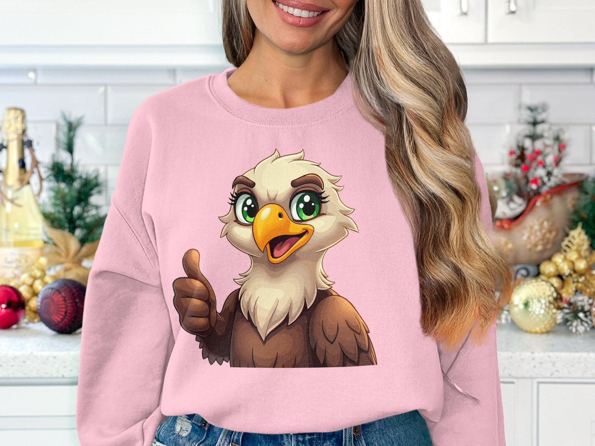 A woman stands in a festive kitchen wearing a pink unisex sweatshirt by Garment Graphics. The design features a cartoon eagle giving a thumbs-up, surrounded by holiday decorations and greenery.