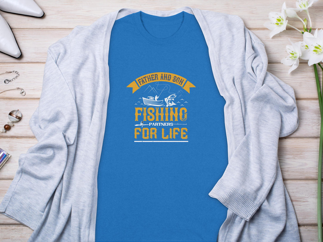 A black Garment Graphics Fishing T-Shirt displays Father and Son Fishing Partners for Life in yellow and white with a fish, rod, and boat design. Its paired with shorts, a basketball, towel, and water bottle on a brown surface—a perfect family bonding scene.