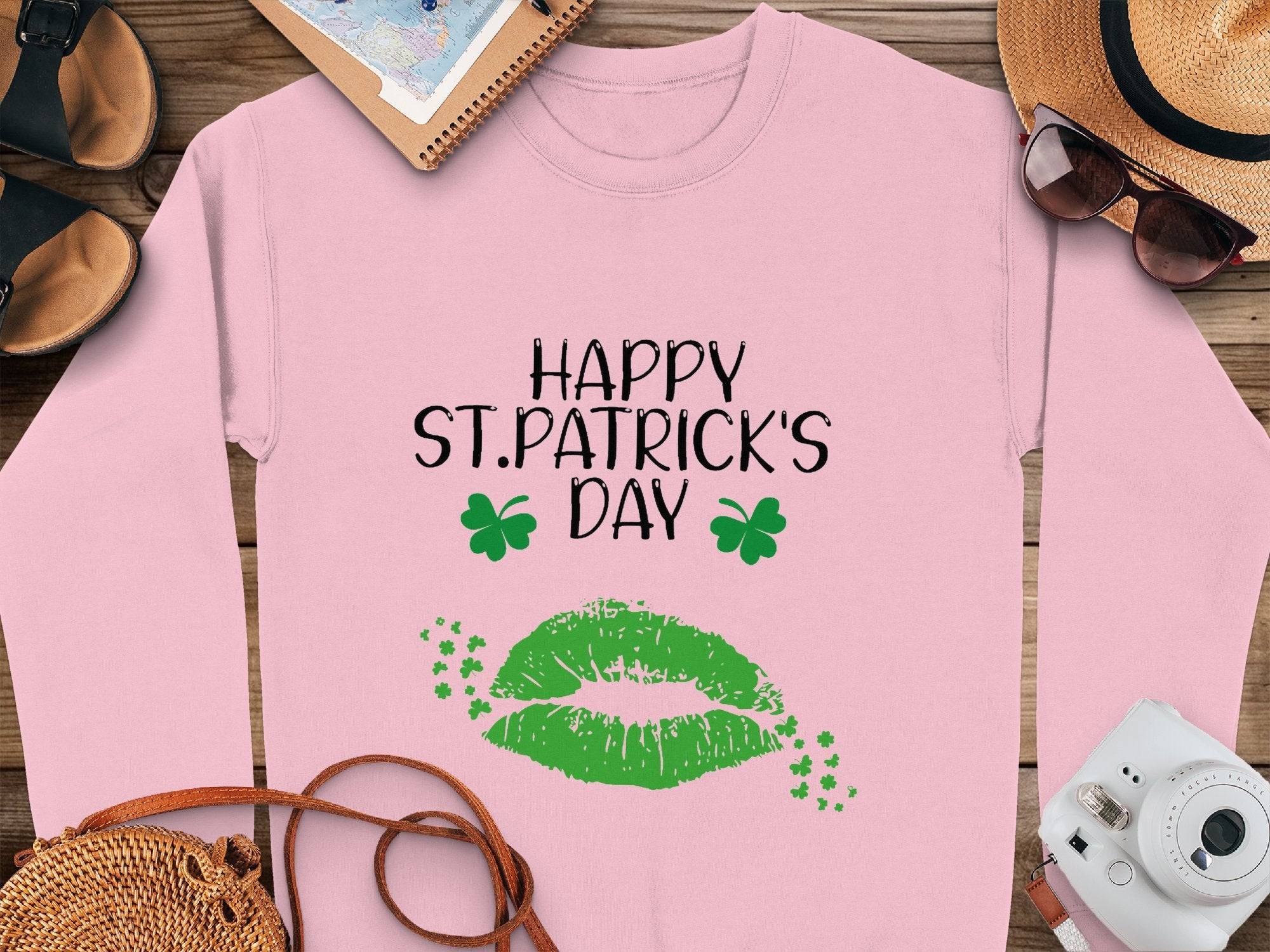 A pink shirt from Garment Graphics radiates festive spirit with Happy St. Patricks Day in black text, accompanied by two green shamrocks and a bold clover kiss design featuring a large green lip print surrounded by clovers, displayed on a table with hat, sunglasses, notebook, and camera.