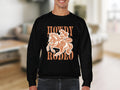 A person wears a Garment Graphics black graphic sweatshirt featuring a cowboy riding a horse with Howdy Rodeo in bold orange letters. This unisex piece stands out against the blurred indoor backdrop, seamlessly blending style and comfort.