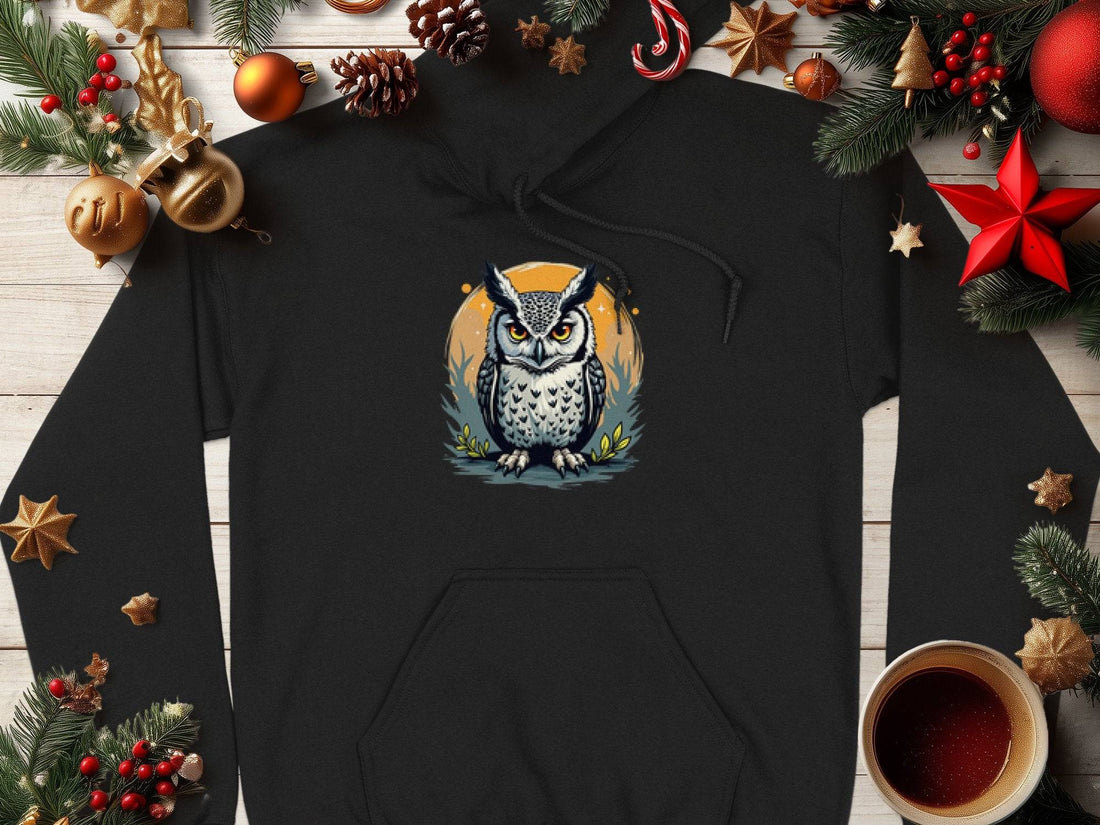 A Garment Graphics black owl artwork hoodie lies on a white wooden surface, accompanied by festive pine cones, red and gold ornaments, greenery, stars, and a coffee cup. This classic fit hoodie offers enchanting seasonal style for cozy days.