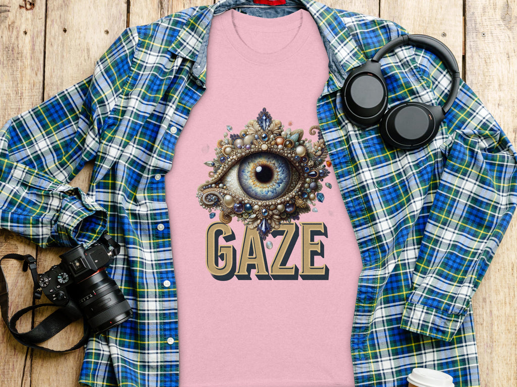 A pink graphic T-shirt by Garment Graphics, featuring an intricate eye gaze design and the word GAZE, lies on a wooden surface. Nearby are a blue classic fit plaid shirt, a DSLR camera, headphones, and a white coffee cup.