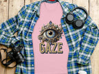 A pink graphic T-shirt by Garment Graphics, featuring an intricate eye gaze design and the word GAZE, lies on a wooden surface. Nearby are a blue classic fit plaid shirt, a DSLR camera, headphones, and a white coffee cup.