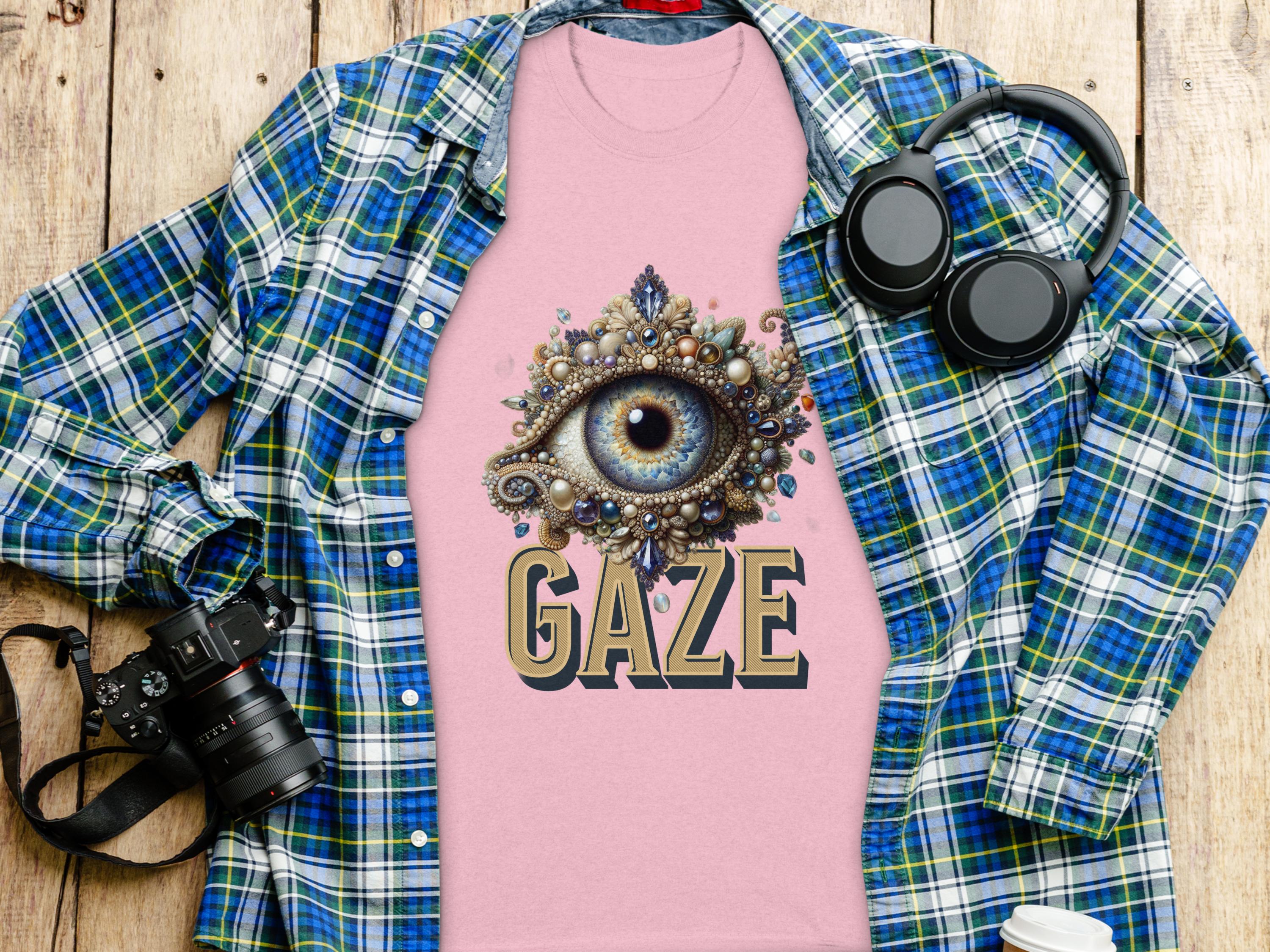 A pink graphic T-shirt by Garment Graphics, featuring an intricate eye gaze design and the word GAZE, lies on a wooden surface. Nearby are a blue classic fit plaid shirt, a DSLR camera, headphones, and a white coffee cup.