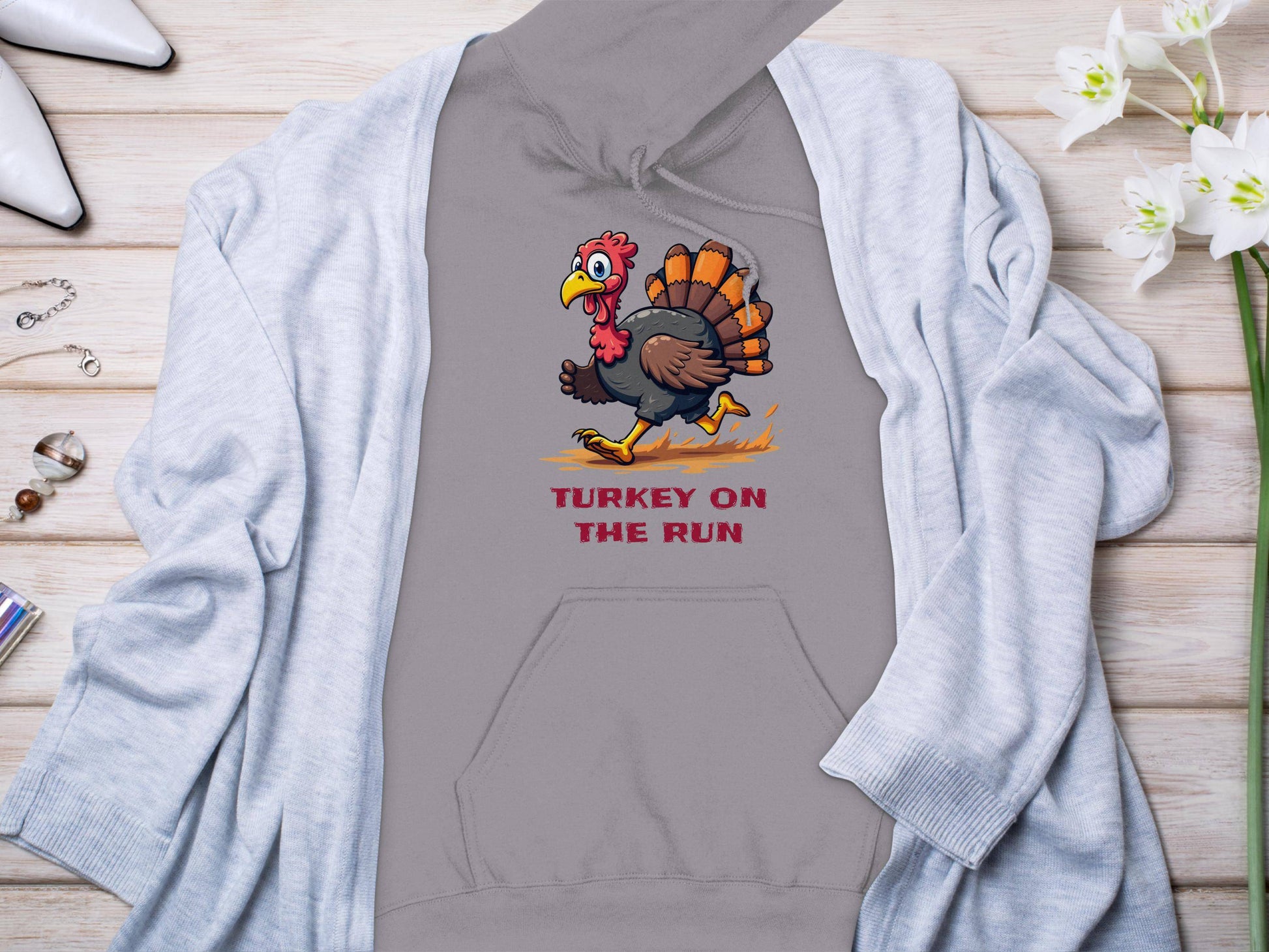 A Garment Graphics gray hoodie with a cartoon turkey and Turkey On The Run is stylishly paired with a light gray cardigan on a wooden floor, accented by accessories and white flowers—ideal for fans of humorous hoodies.