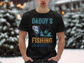 A person stands outdoors in a Garment Graphics t-shirt featuring Daddys Fishing Buddy with a boat and fish design, ideal for fishing enthusiasts. Blurred snow-covered trees create a serene backdrop to this fun tribute to fishing adventures.