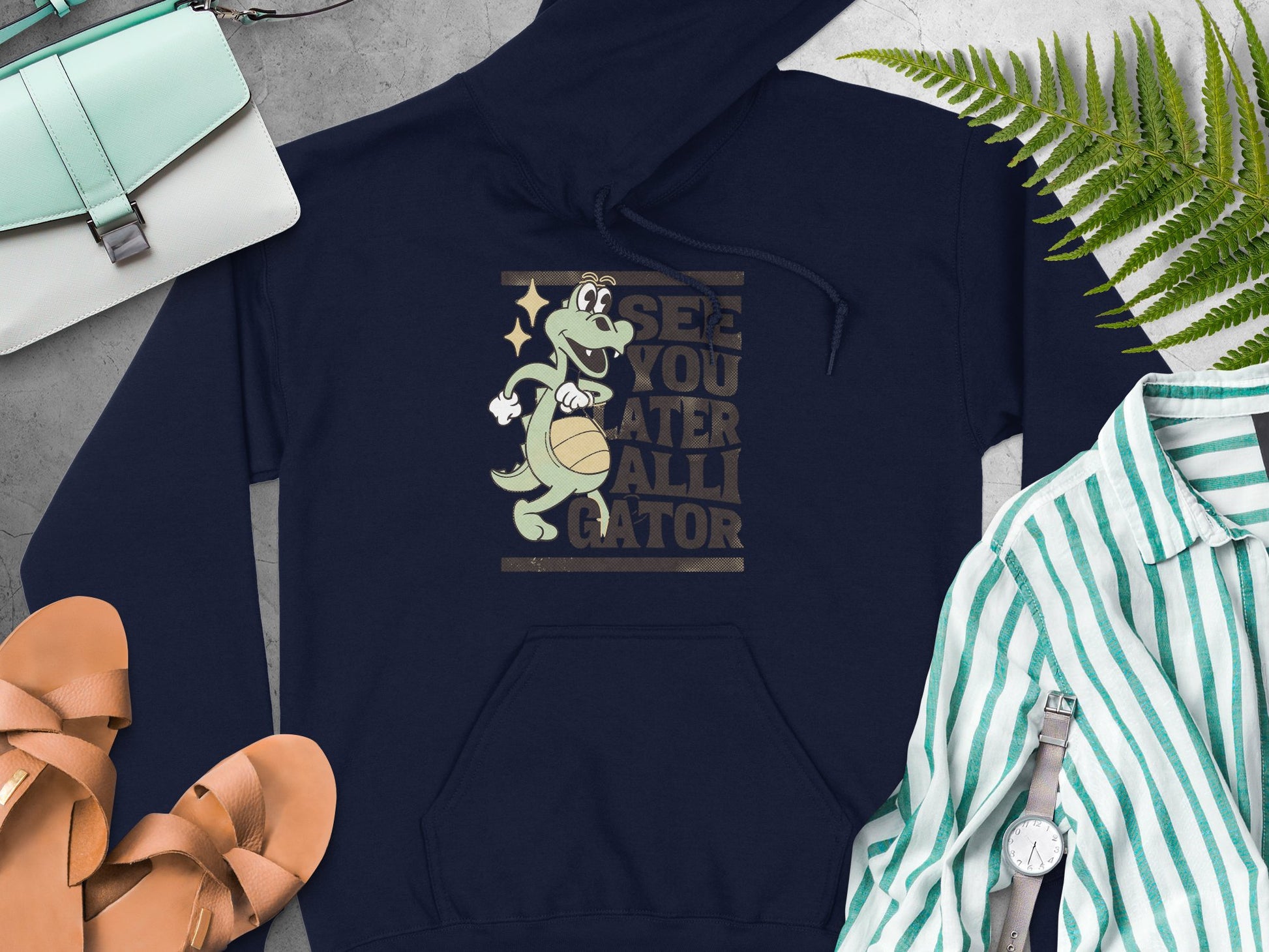 A navy blue See You Later Alligator hoodie from Garment Graphics features a gator graphic with a basketball. Its surrounded by a green purse, tan sandals, a striped shirt, watch, and green fern leaves on a gray surface.