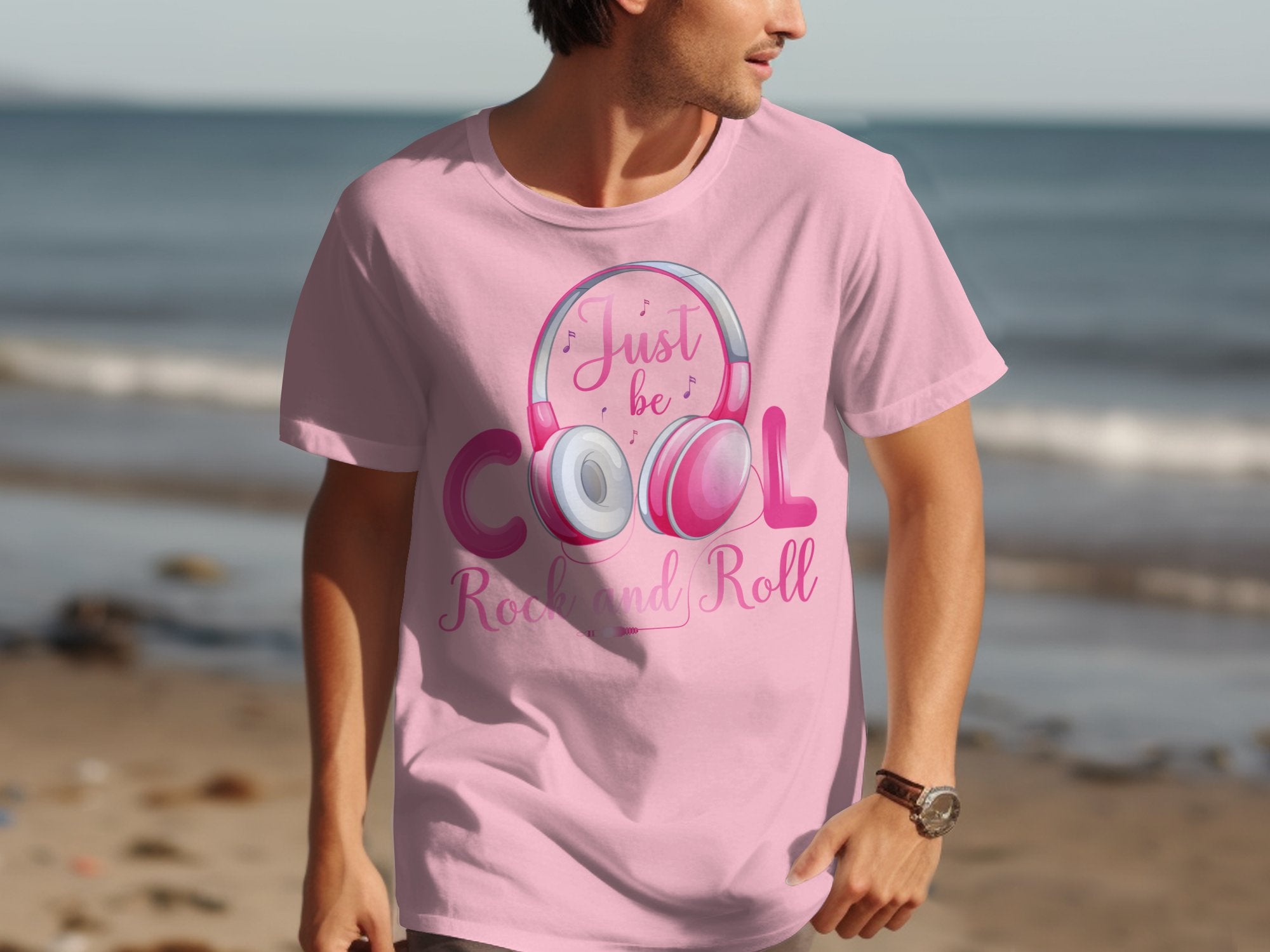 A man wearing a Garment Graphics light pink t-shirt featuring the slogan Just be Cool, Rock and Roll for music lovers walks along a beach with fun headphones, gazing to his right against waves in the background.