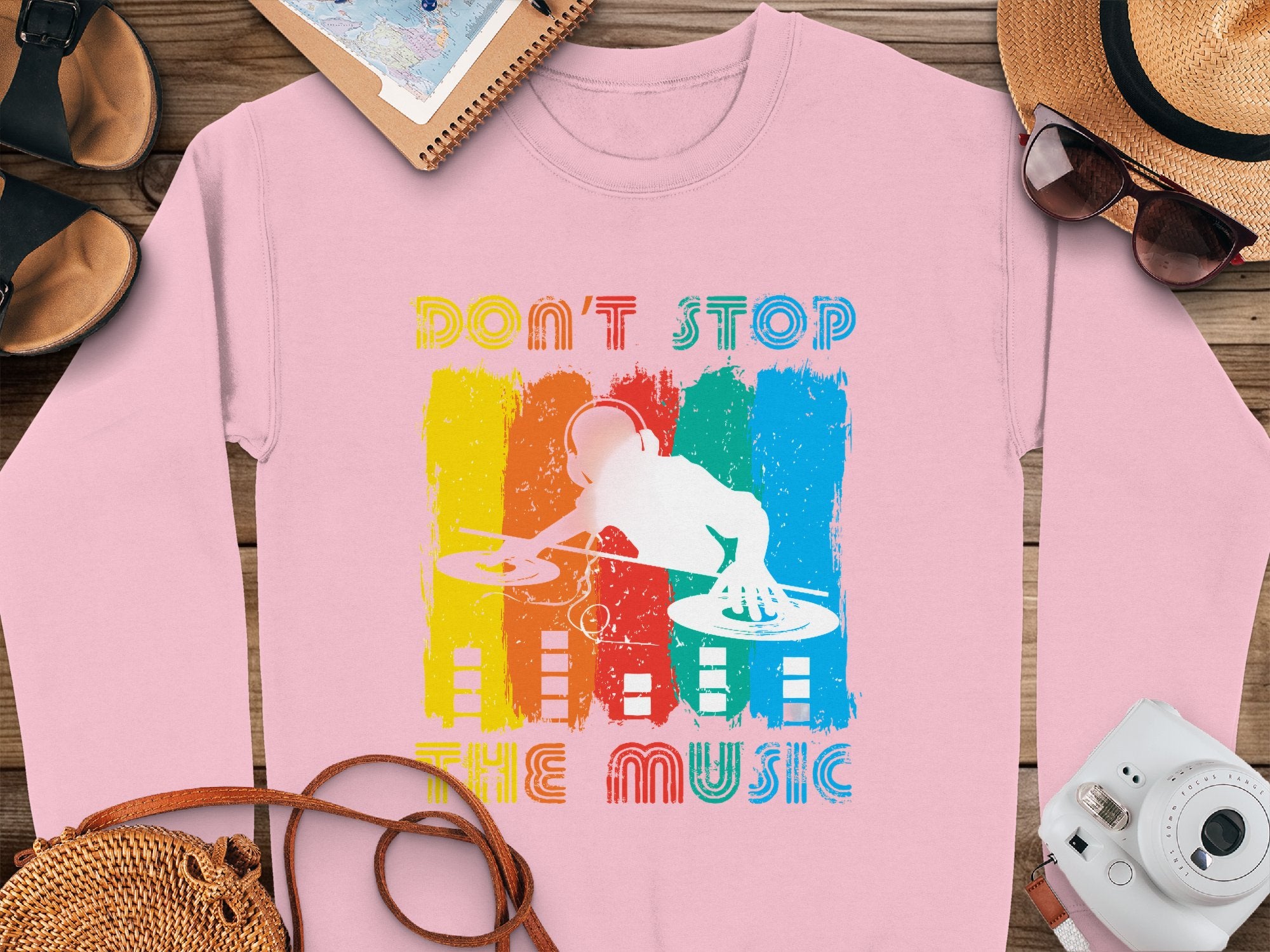 A pink sweatshirt by Garment Graphics featuring a DJ mixing on turntables over a rainbow paint splatter. Perfect for music fans, it reads Dont Stop the Music. Displayed with a hat, sunglasses, shoes, and notebook on a wooden surface.