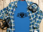 Flat lay of a blue graphic t-shirt featuring SCANIA POWER and an eagle logo by Garment Graphics, artfully arranged on a wooden surface. The stylish setup includes a blue plaid shirt, black headphones, camera, and coffee cup lid.