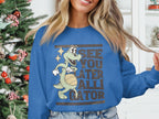 A person wears a Garment Graphics blue sweatshirt with an alligator graphic holding a basketball and the phrase See You Later Alligator, amidst a festive Christmas tree backdrop.
