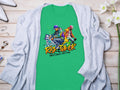 A Garment Graphics green t-shirt with Risk Taker 10th Final T-Shirts and two street dancers graphic captures street dance culture. Laid on wood, paired with a light gray cardigan, with white flowers and a perfume bottle creating an artistic setup.