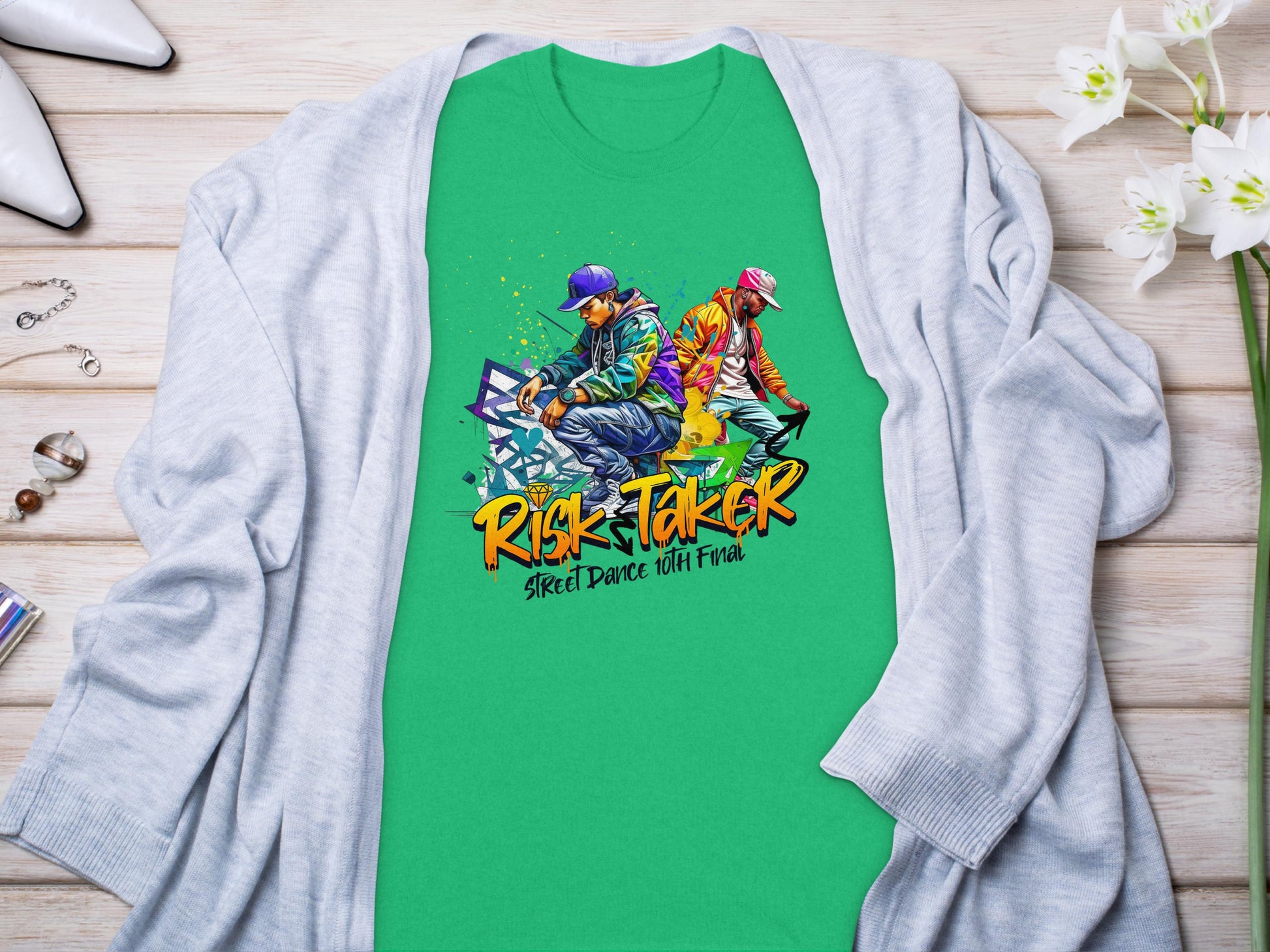 A Garment Graphics green t-shirt with Risk Taker 10th Final T-Shirts and two street dancers graphic captures street dance culture. Laid on wood, paired with a light gray cardigan, with white flowers and a perfume bottle creating an artistic setup.