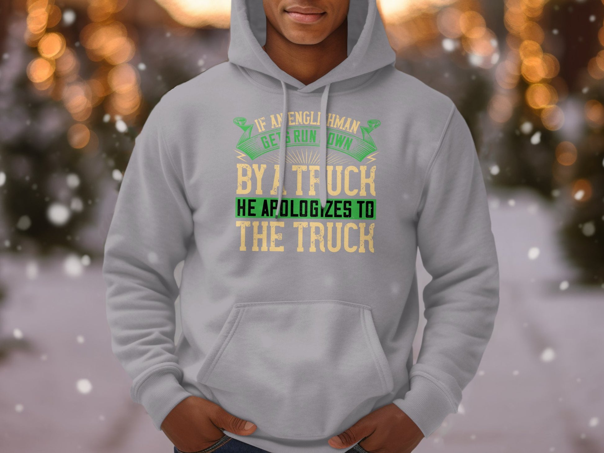 Someone is wearing a Garment Graphics light gray hoodie featuring the witty quote, If an Englishman gets run over by a truck, he apologizes to the truck, against a blurred snowy scene with trees in the background.