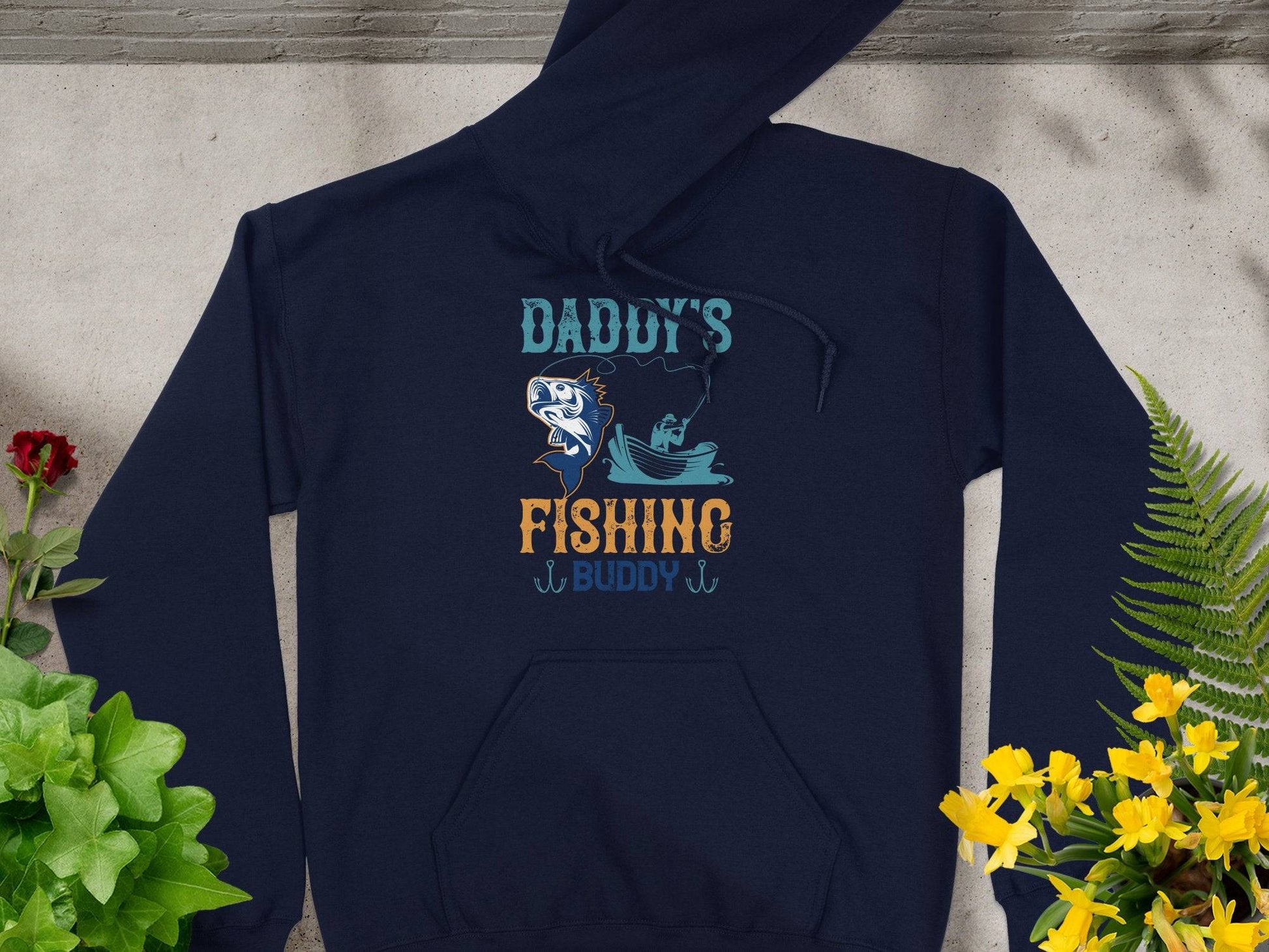 A Garment Graphics navy hoodie lies on concrete amid vibrant plants and flowers, perfect for outdoor adventures. It features a fish and boat design with Daddys Fishing Buddy in bold blue and orange text.