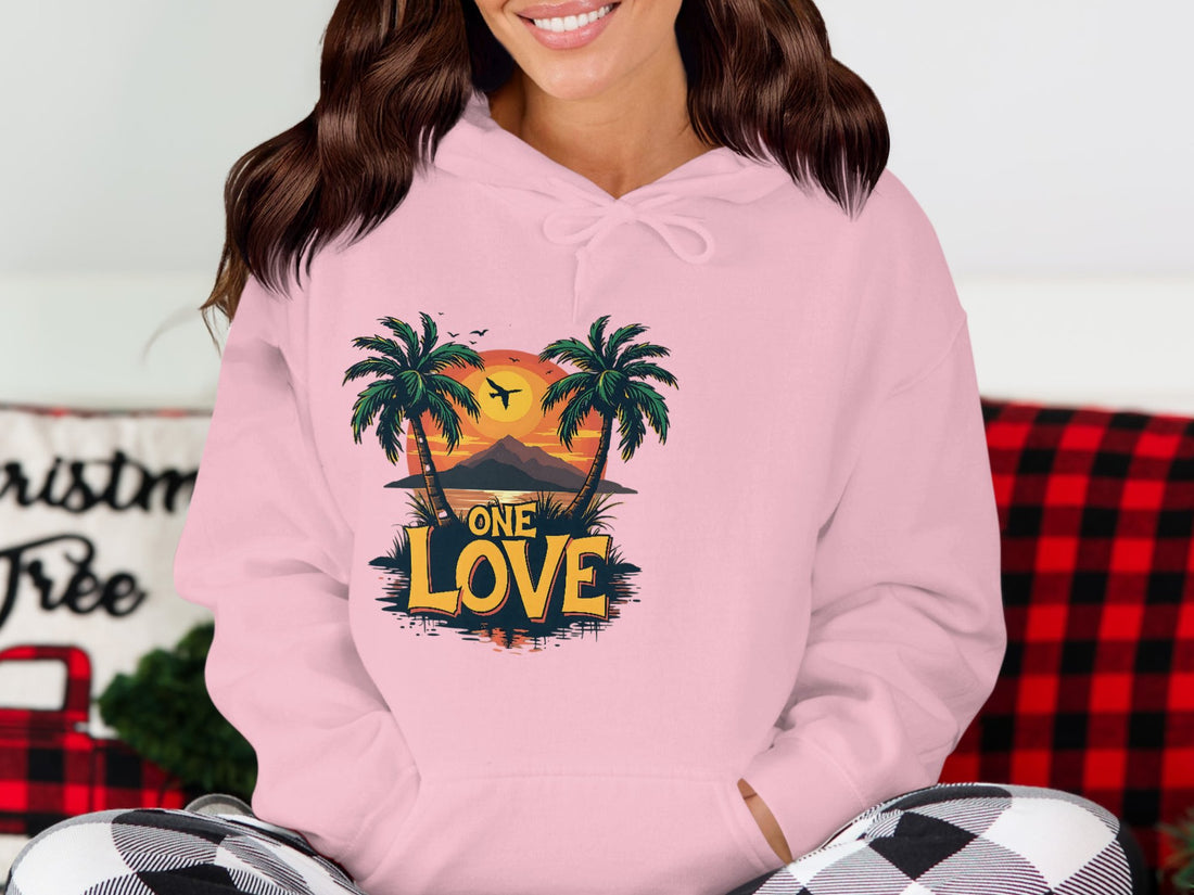 Wearing Garment Graphics pink hoodie featuring a tropical beach with palm trees, vibrant sunset, and ONE LOVE, a person embraces their style while sitting comfortably with a plaid blanket in the background.