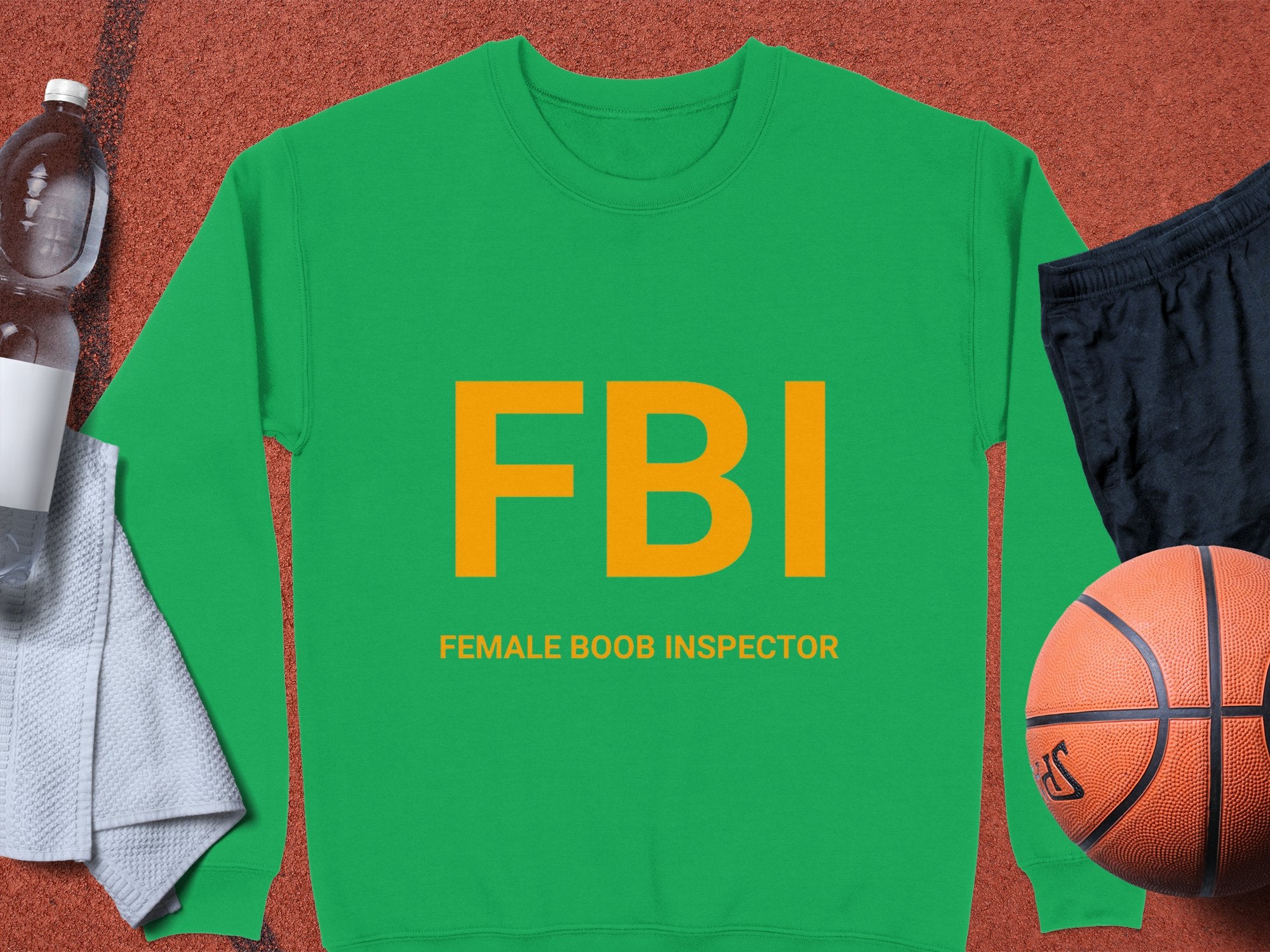 This unisex Garment Graphics green sweatshirt boasts bold yellow FBI lettering with Female Boob Inspector beneath. Its shown on a brown surface with a white bottle, gray towel, black shorts, and basketball. Ideal for those who appreciate humor and style!.