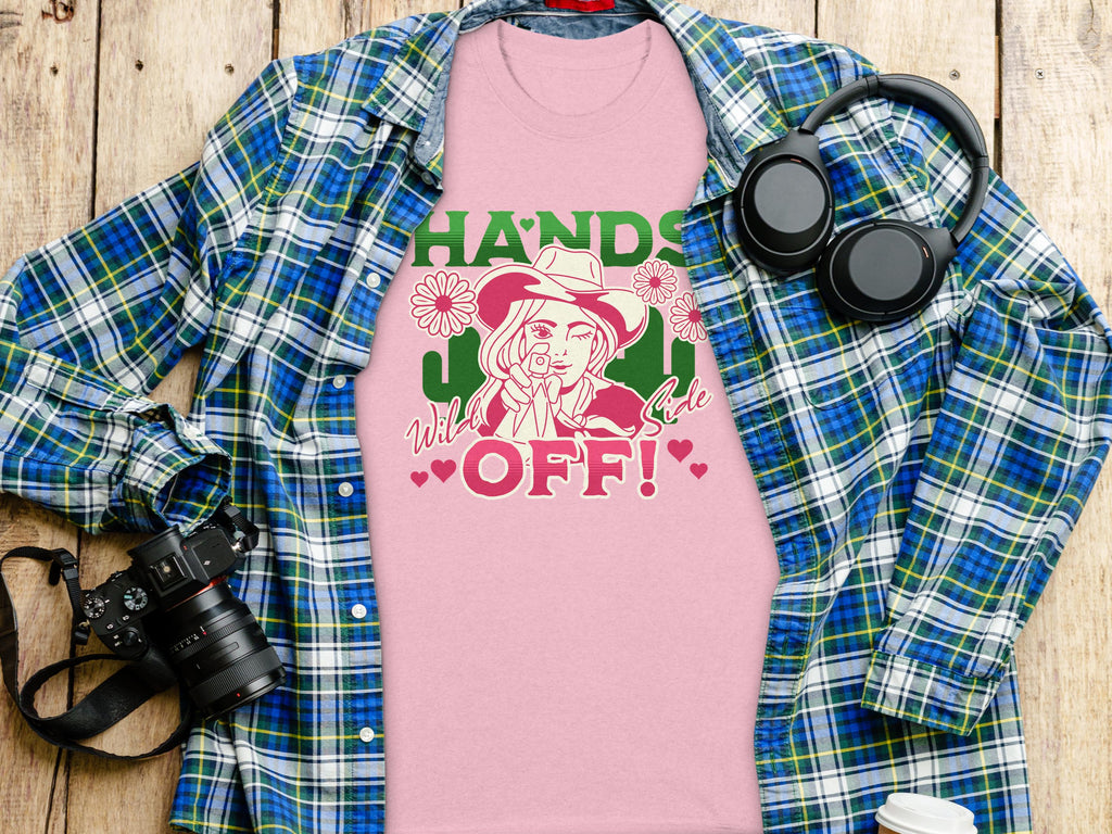Garment Graphics presents a pink T-shirt with a bold Hands Off! floral motif and cowboy design, featuring a classic fit. Its elegantly displayed on wood beside a blue-green plaid shirt, headphones, white water bottle, and black camera.